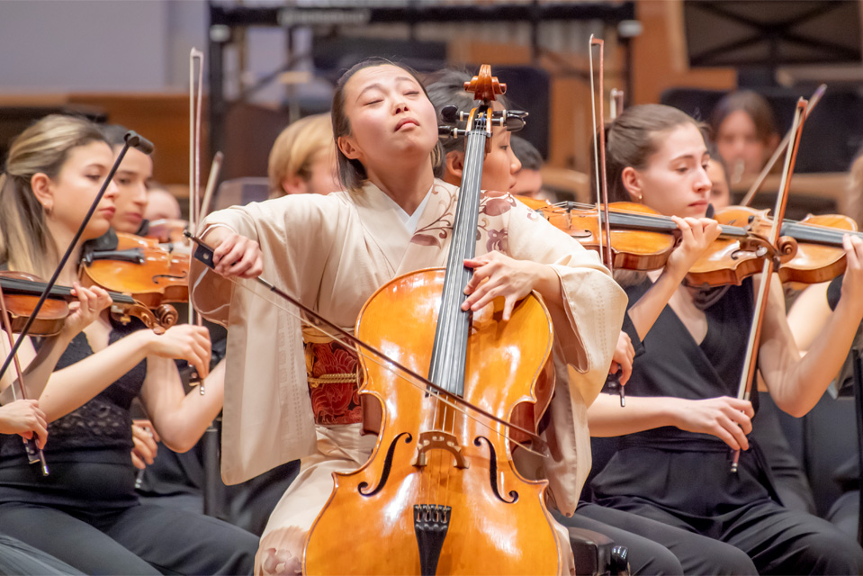 An Asian female student, wearing a formal kimono, playing the cello, with students performing behind her in an orchestra.